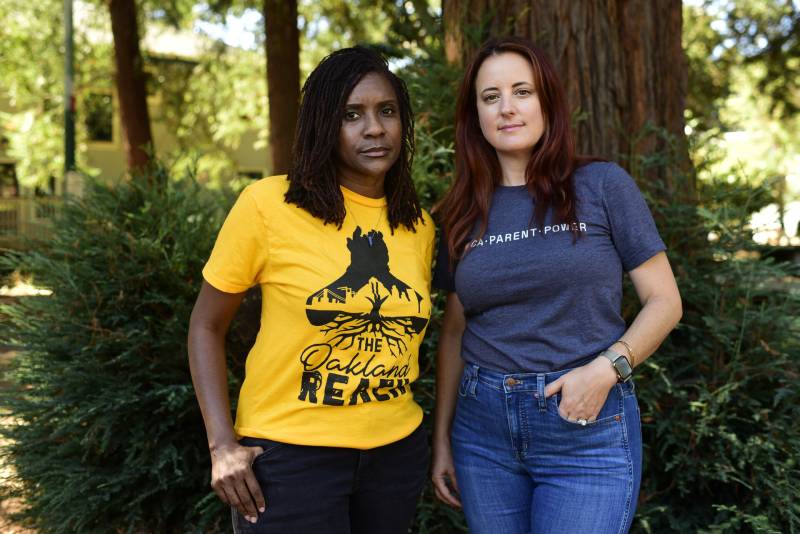 Two women are standing in woods. The woman on the left is wearing a yellow t-shirt and the woman on the right is wearing a navy blue t-shirt.
