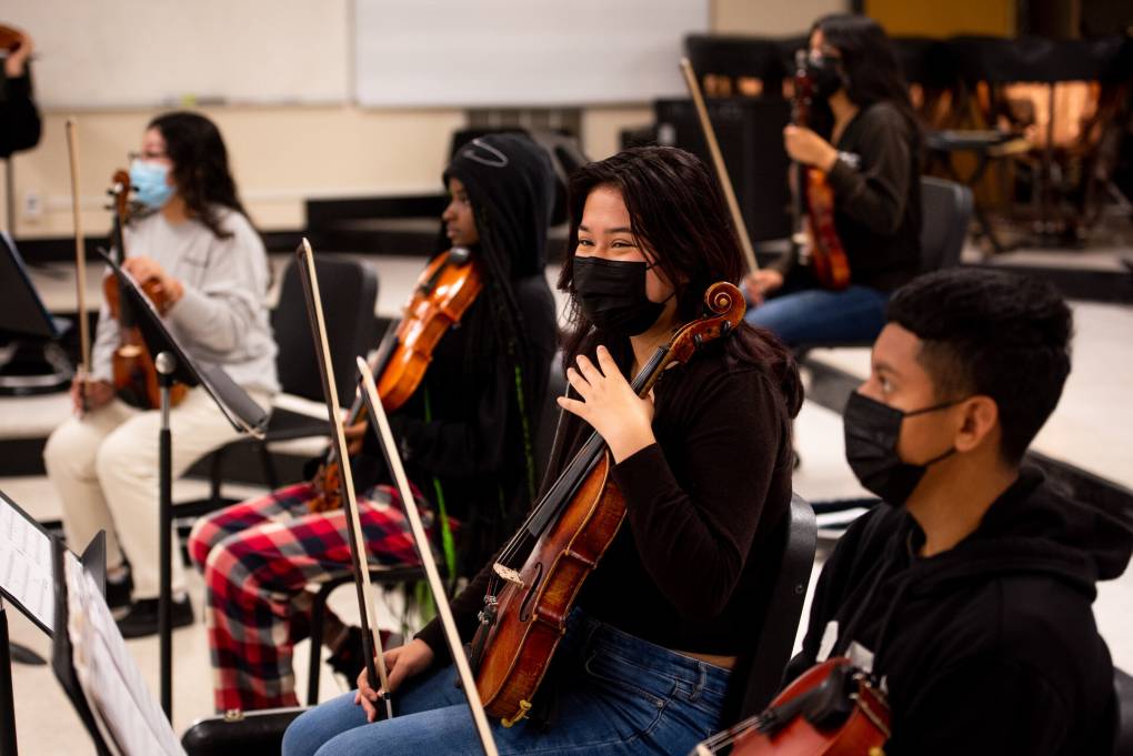Students in the school orchestra sit facing their teacher, a black girl on violin, a Latina girl on viola, and a Latino boy on violin.