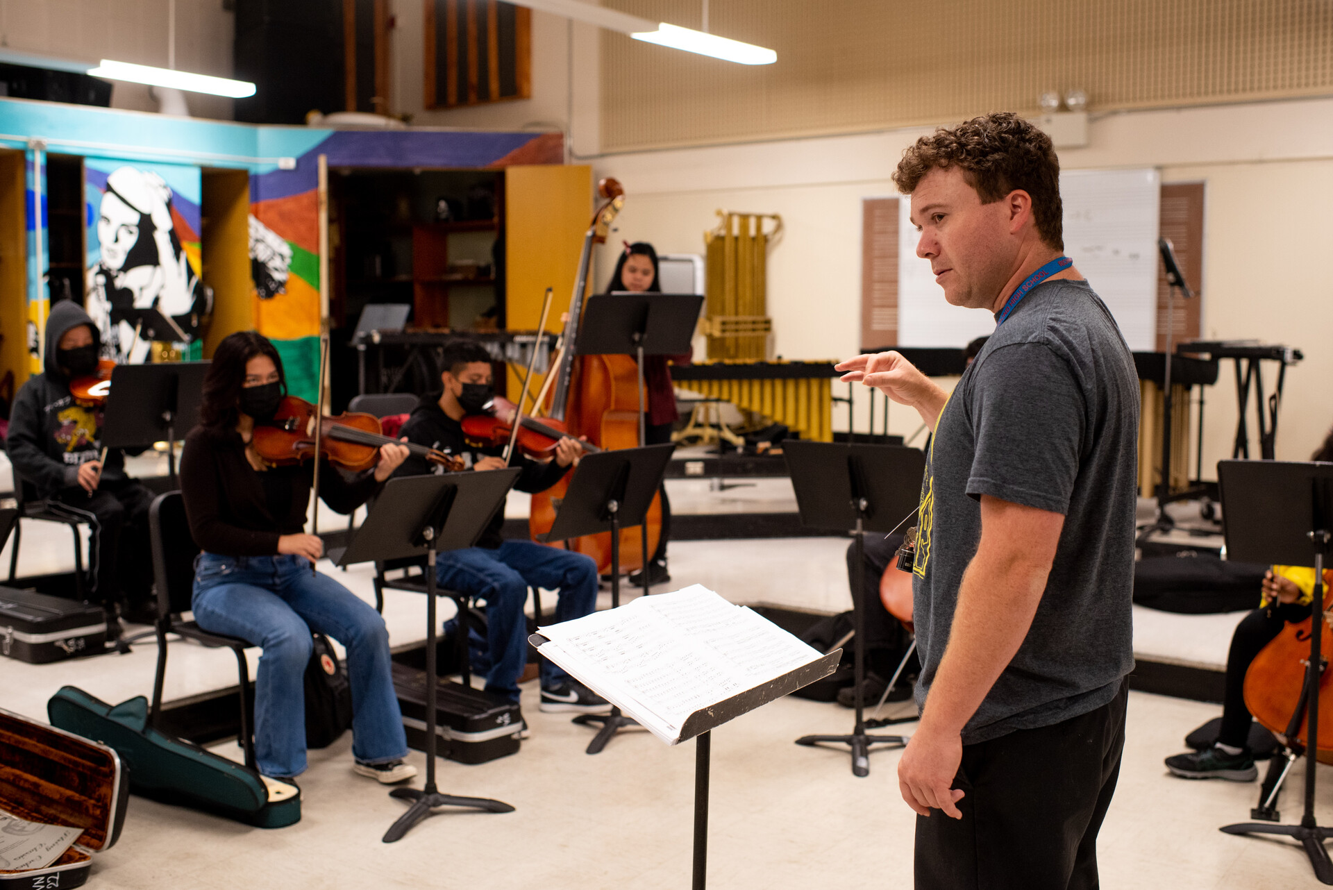 A white music teacher in his 30s teaches a small orchestra of kids playing various instruments in a high school music classroom.