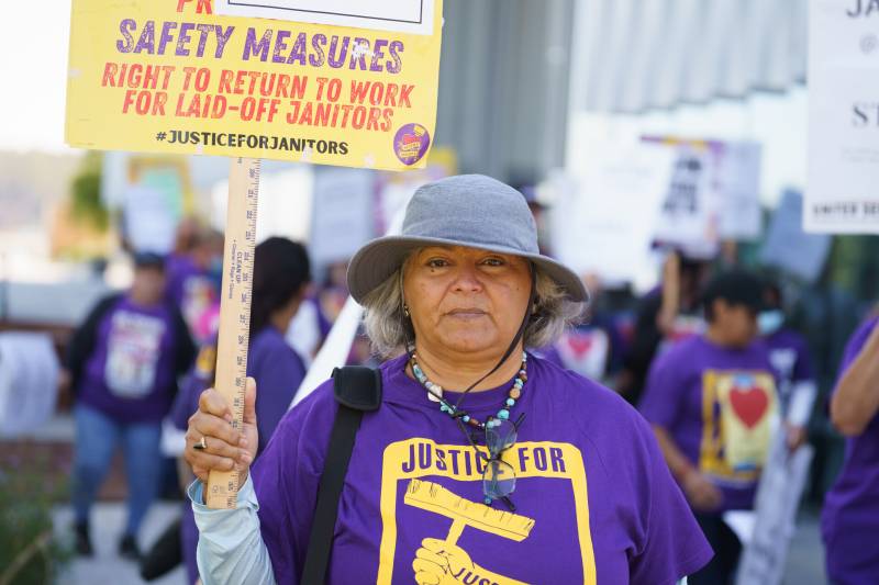 A person wearing a grey wide-brimmed hat and purple shirt reading justice for janitors faces the camera while holding a picket sign.