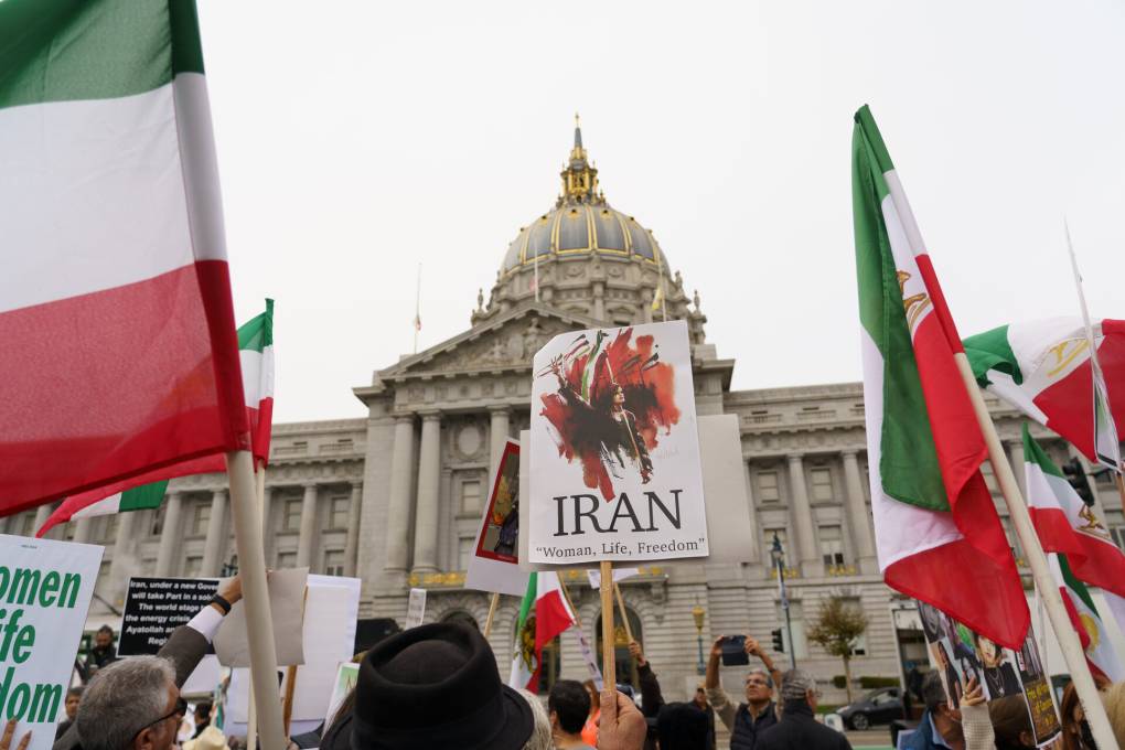 Several people fly Iranian flags and homemade signs in front of a building.