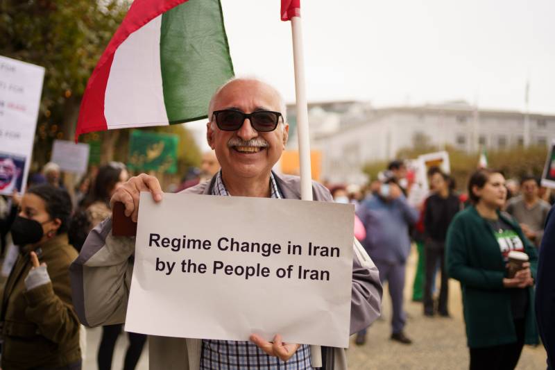 A man wearing sunglasses holds a sign that says "Regime Change in Iran by the People of Iran" in a crowd.