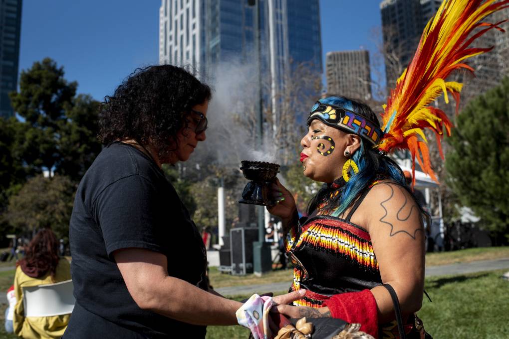 Two people stand facing each other in the middle of a park setting with tall city buildings behind them. The person on the right is dressed in Indigenous regalia, and is blowing a smoky substance from a dish held in their right hand. Their other hand is holding the arm of the person on the left.