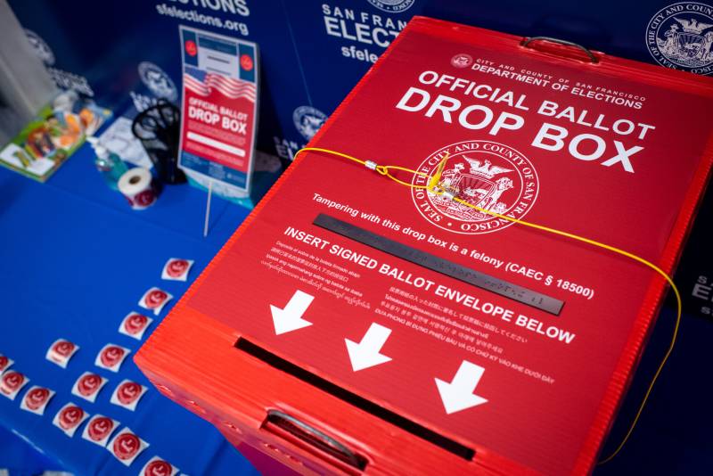Bright red box reading 'official ballot drop box' on a bright blue table