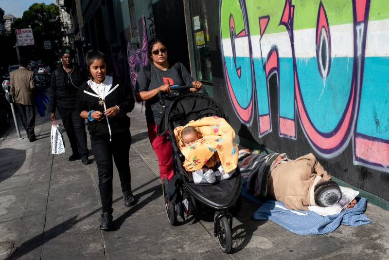 A woman standing next to a girl pushes a stroller with a baby wrapped in a yellow blanket past a person lying down on the sidewalk.