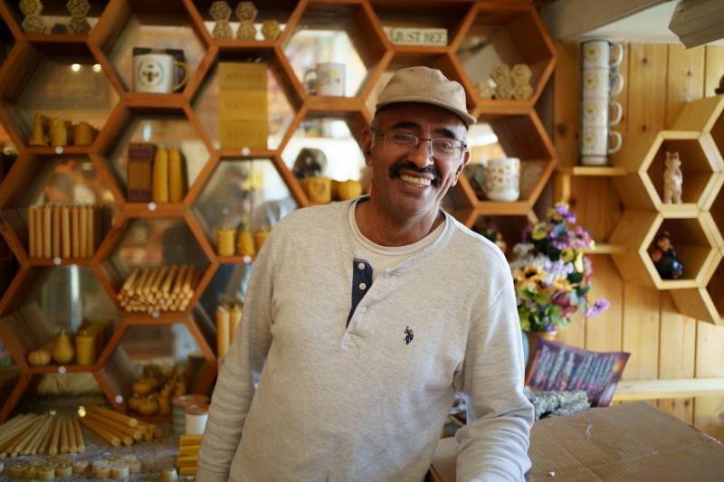 a smiling Middle Eastern man in a white shirt stands in front of a honeycomb-shaped shelf