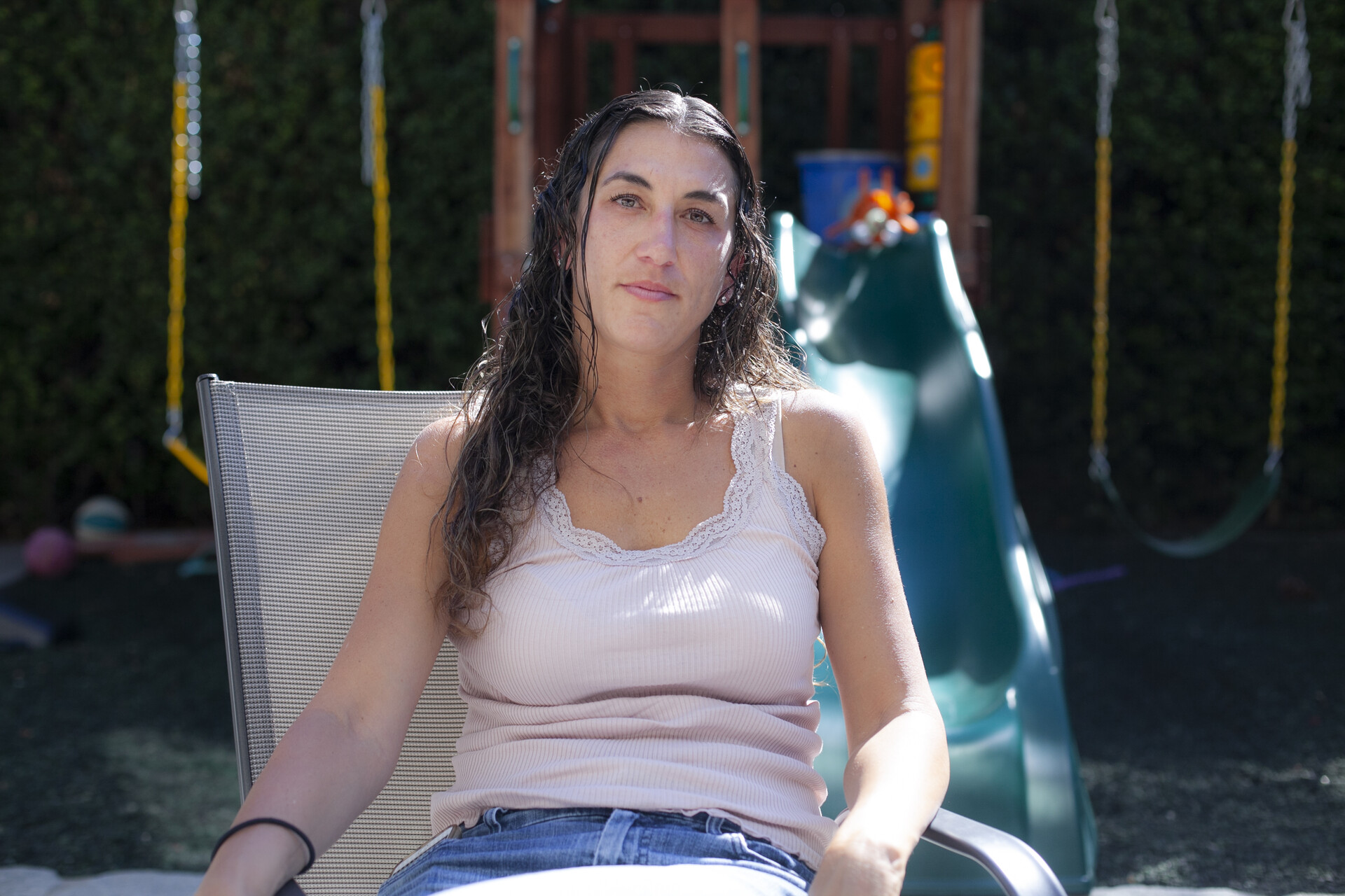 A woman wearing a light tank top shirt and blue jeans sits in a chair outside.