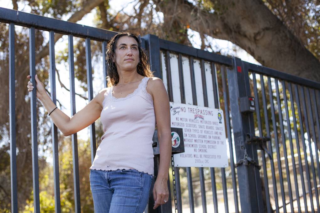 A woman wearing a light top and blue jeans holds on to a gate outside with one hand.