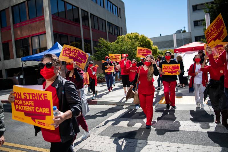 People holding yellow and red signs and wearing red nurse outfits walk across the street.
