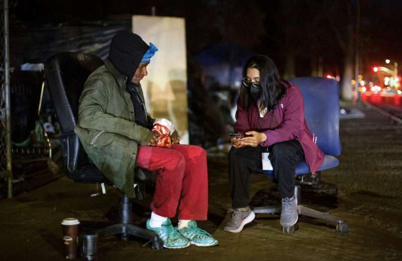 Two people sitting outside. The person on the left is wearing a green jacket, black hoodie and red pants. The person on the right is wearing a purple coat, black mask and black pants.