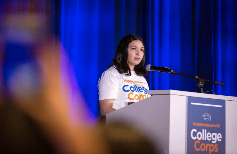A woman stands at a podium wearing a white t-shirt that says "College Corps."