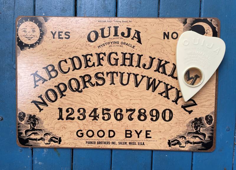 A Ouija board featuring the alphabet, numbers zero through nine, yes, no, and goodbye. It sits on a blue wooden deck, with the white planchette placed on the board over the letter M.