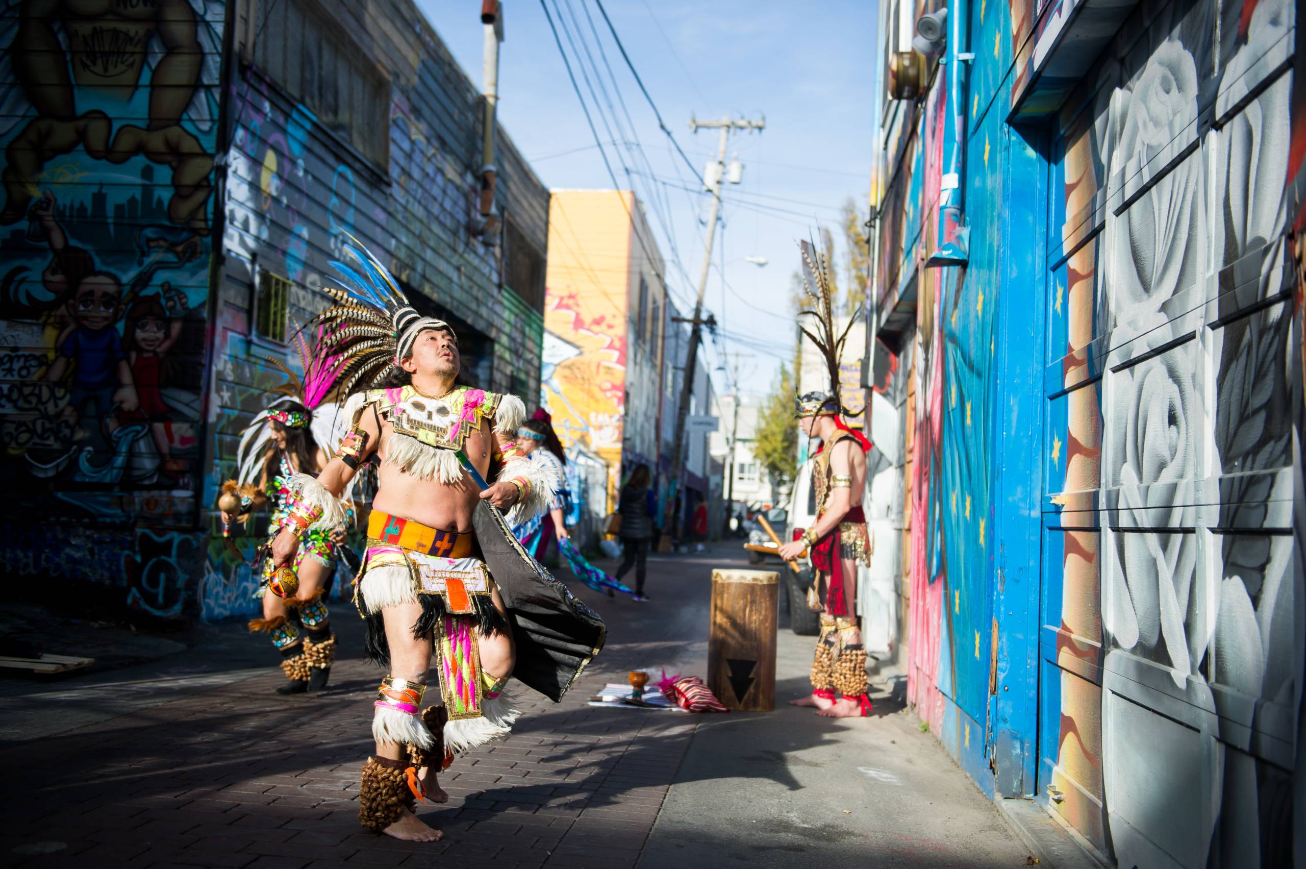 Aztec dancer in traditional dresswith head raised dances with others in a Mission District alley surrounded by colorful murals