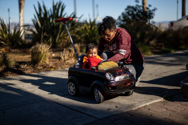 A woman wearing glasses puts a small child into a stroller shaped like a toy car.