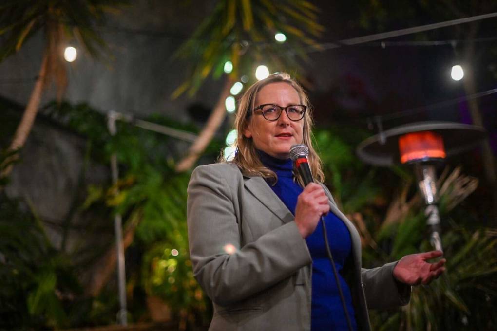 A white woman with a grey suit over a blue sweater speaks into a microphone at an event