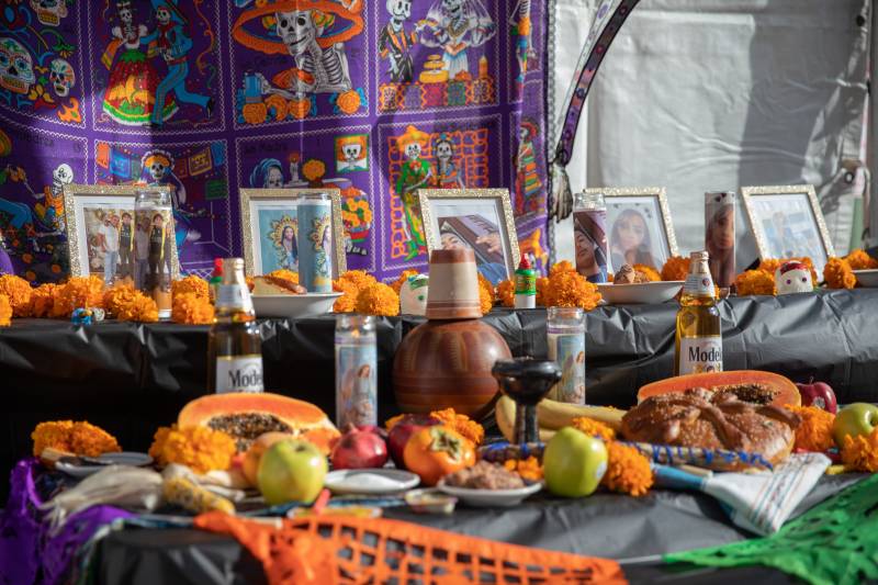 Framed photos of deceased loved ones sit behind tall candles also printed with the faces of the deceased. The alter is adorned with offerings from the dead, including fruits, bread, candy and beer. Everything sits on a black table cloth decorated with marigolds.