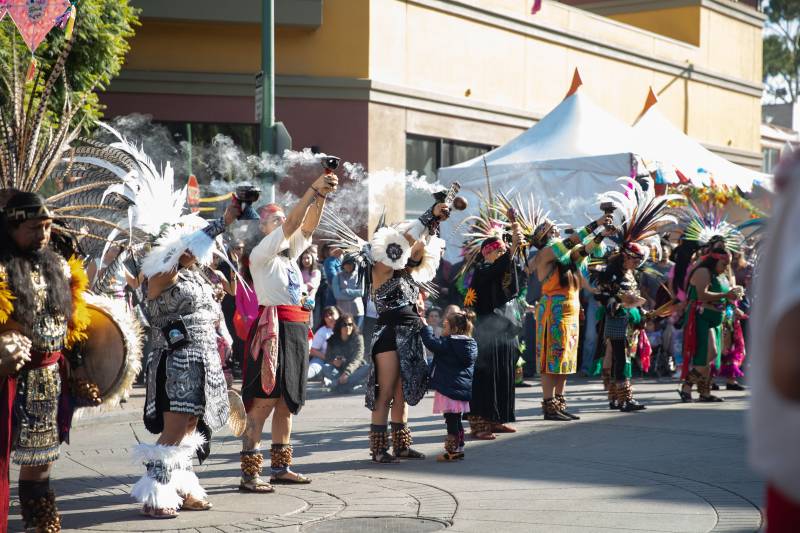 A group of dancers wearing various styles of traditional clothing, several including feathered headpieces, stand in a line side by side. Their arms extended, most appear to hold goblets with smoke coming from them. The performers stand amid a larger crowd of onlookers. 