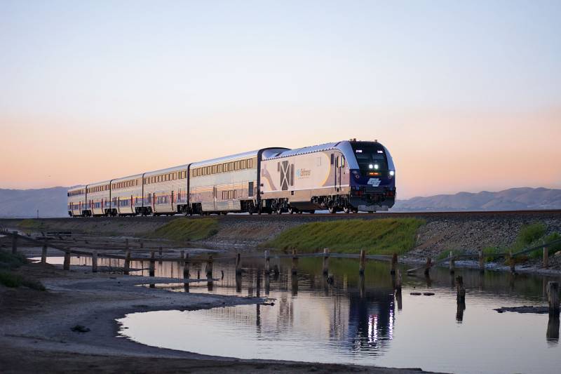 A passenger train reflected in a trackside puddle at sunset.