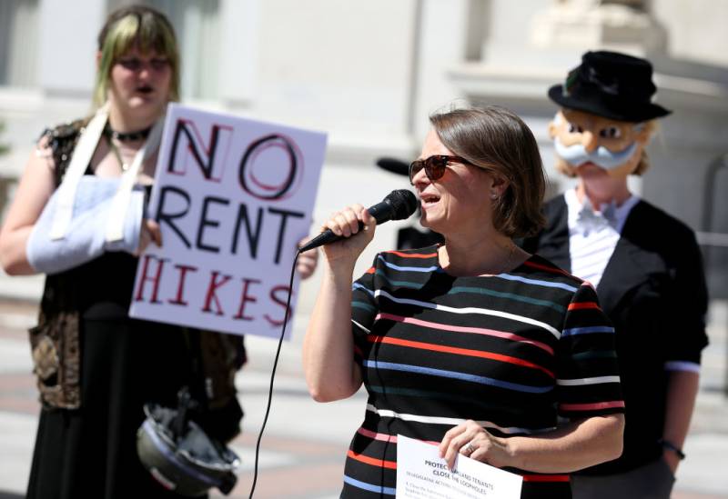 a woman in a striped shirt speaks at a rally while a person holds a sign that says 'no rent hikes'