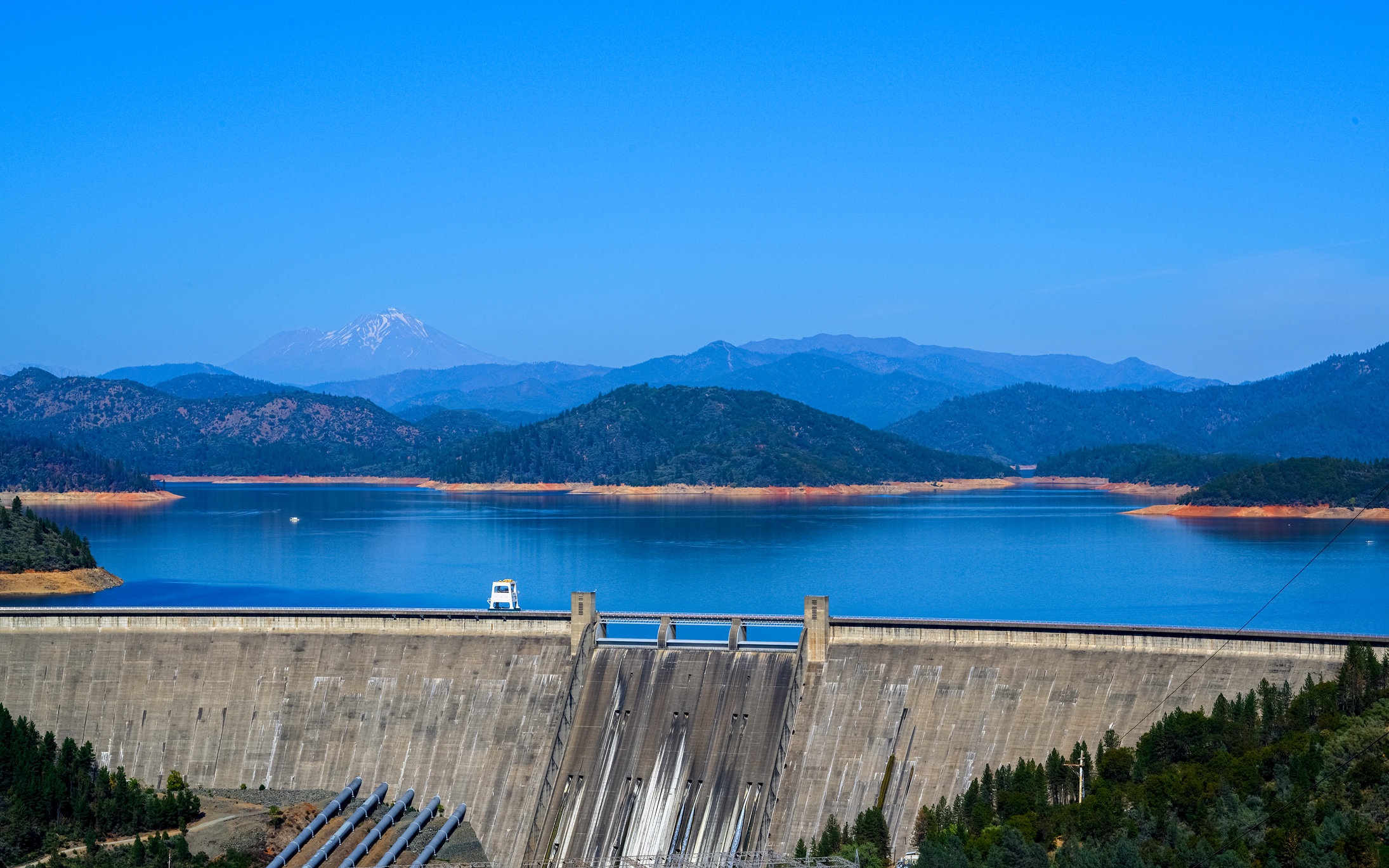 A view of Shasta Dam, a dam wall with big blue reservoir of Shasta lake behind it and treelined hills in the background.