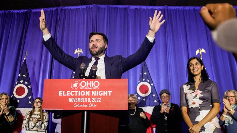 White man in suit speaks with both arms raised high, smiling people around him