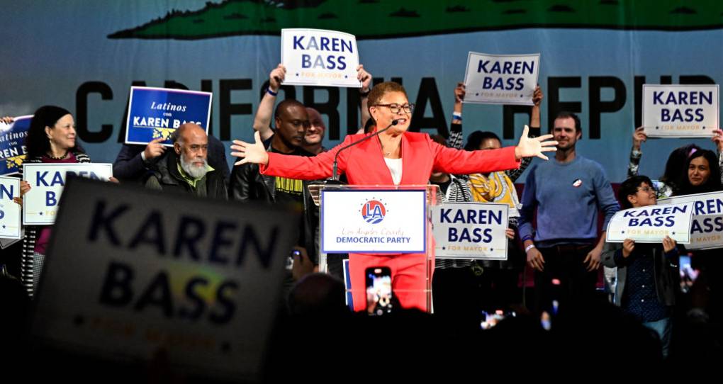 A black woman with a red suit on stage as her supporters rally around her carrying signs that read "Karen Bass"