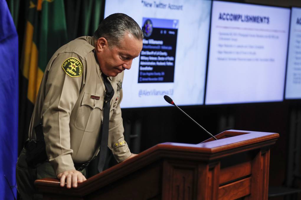 Villanueva stands at a podium, his hands hold the sides of the podium and it appears as though he's bracing himself. The outgoing sheriff looks down, seemingly dejected.