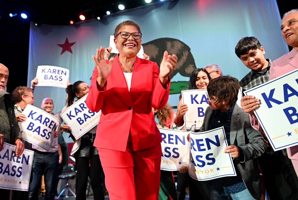 Karen Bass in a red suit is smiling and clapping, surrounded by supporters with her name