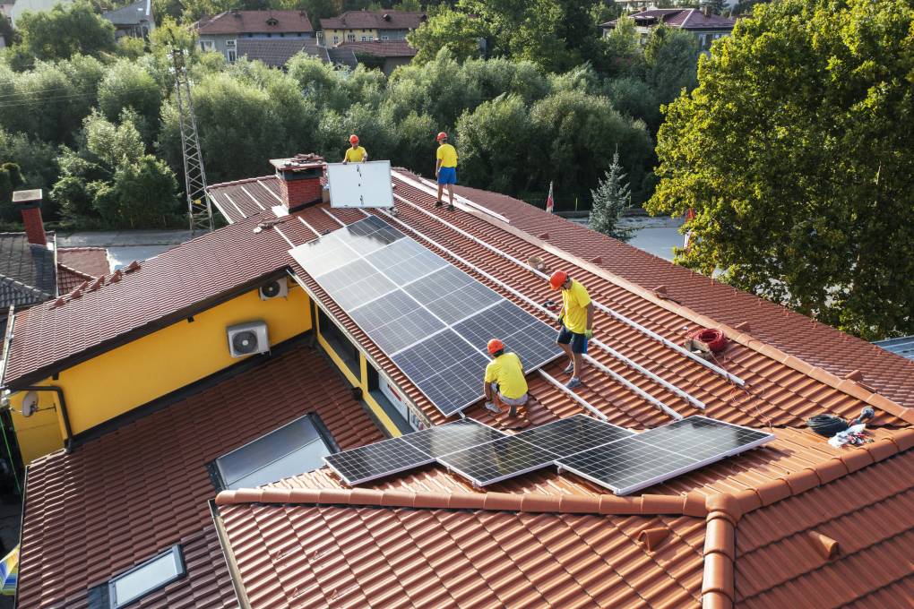 Workers install solar panels on a red rooftop.