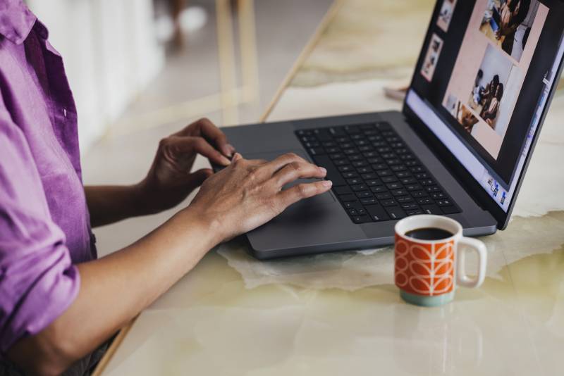 A woman scrolling on her laptop with a coffee mug next to her..