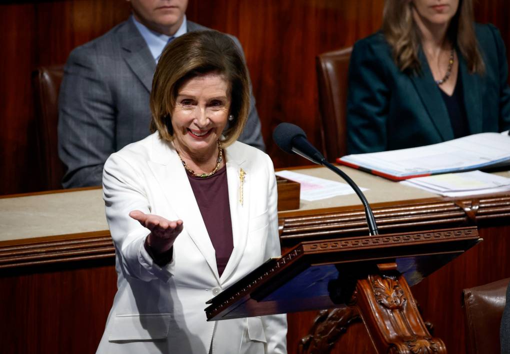 Nancy Pelosi stands in white at a podium at the House, clad in white, smiling, one hand outstretched, palm up.