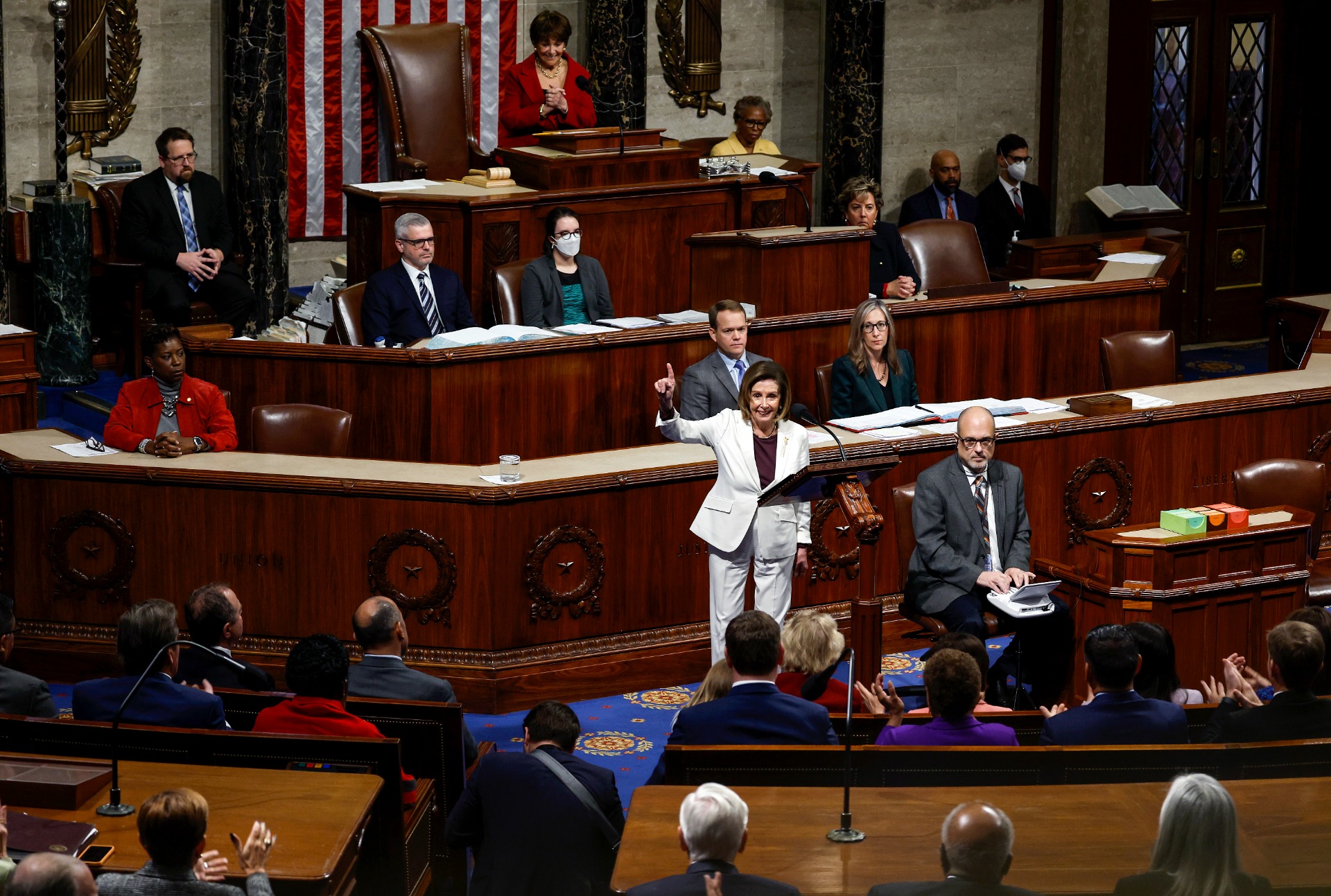 Nancy Pelosi stands on the floor of the House dressed in white, in a long shot showing her colleagues applauding her around her.