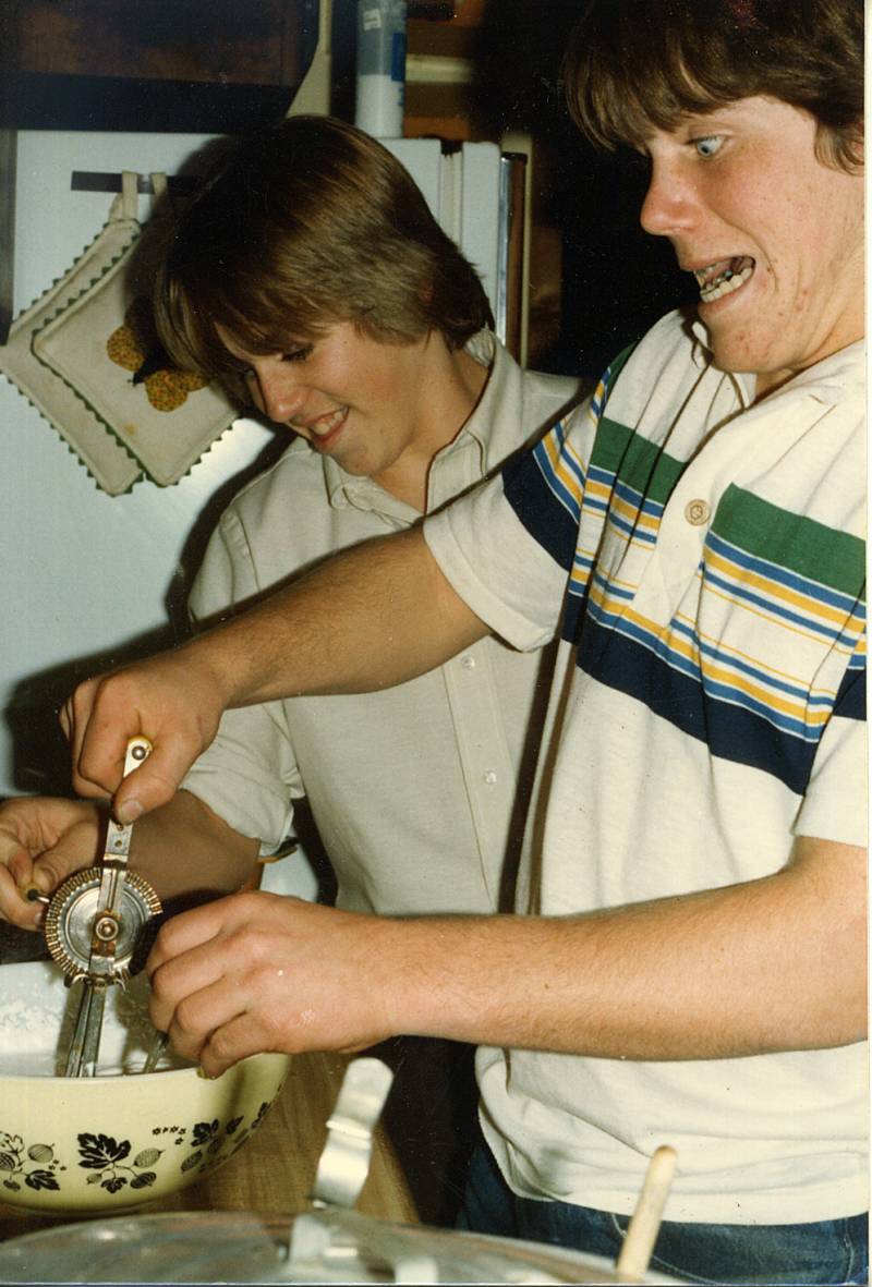 Two young men wearing white shirts in a kitchen with one holding a cooking tool in a bowl.