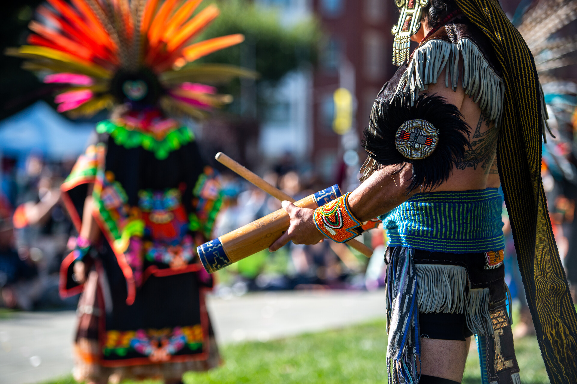 An Aztec dancer in colorful feathered head dresses garb plays a traditional percussion instrument as another dancer stands in the background on a lawn surrounded by onlookers