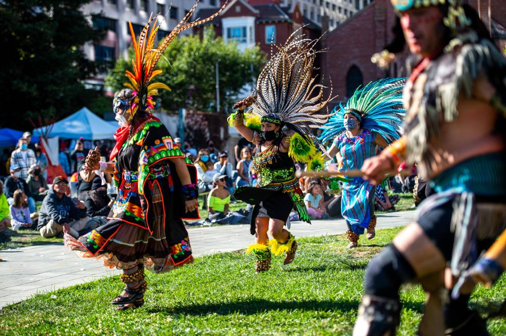 Four Aztec dancers in elaborate colorful garb and feathers dance on a lawn surrounded by onlookers