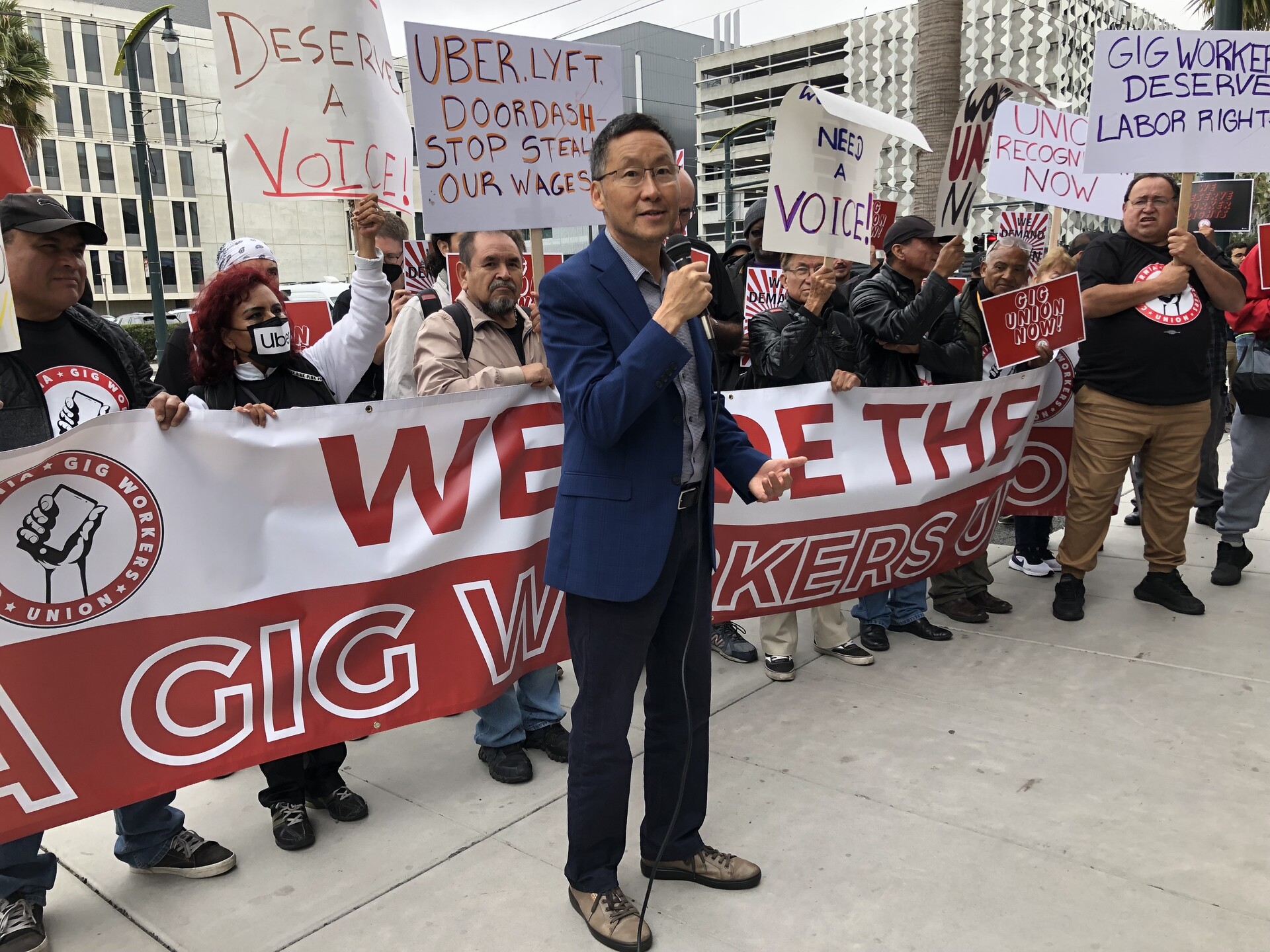 An Asian man in a blue suit holding a microphone and speaking with protesters behind him in support of gig workers.