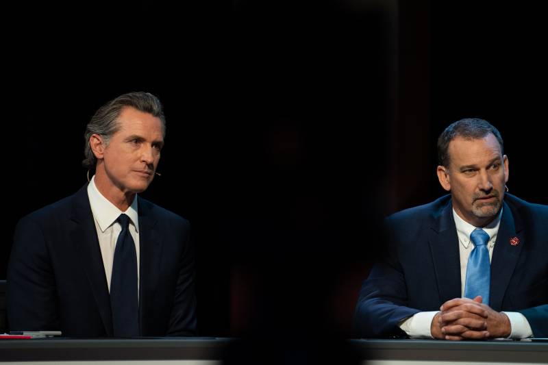 Newsom sits left wearing a dark blue suit and matching tie, Brian Dahle sits right wearing a slightly lighter blue suit and light blue tie. Though the two men are sat next to each other at the same table, an obscured and unidentified object in the center of the foreground divides them.