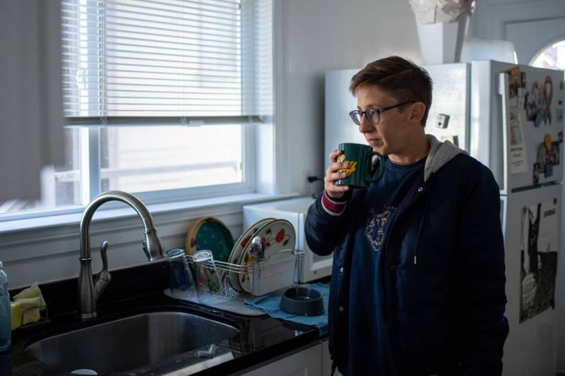 a woman with short hair and glasses stands drinking from a mug by the sink in her apartment kitchen