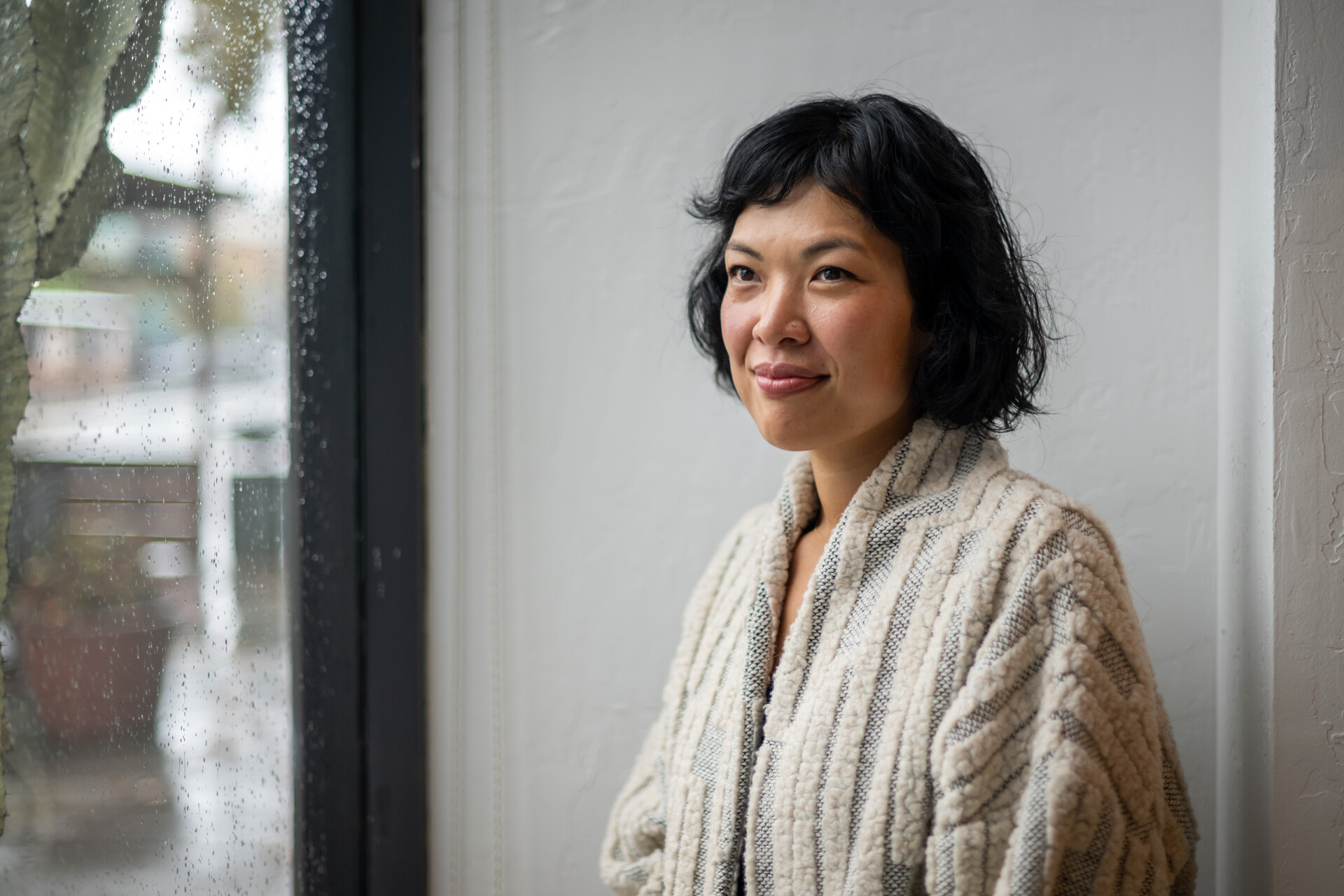 A woman sits right of frame looking left, smiling. She appears to be at a window sill looking out, while wearing a wool cardigan.