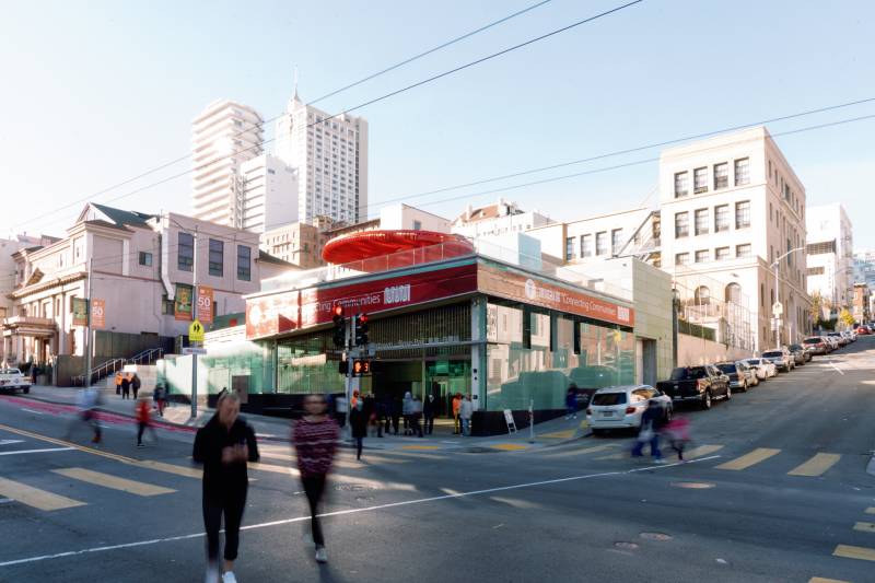 Two people cross the street at a busy intersection with the new Rose Pak SFMTA station on the diagonal corner with a city skyline backdrop.
