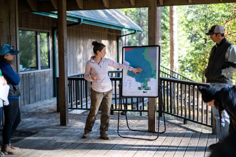 a white woman stands on a deck showing a fire map in a forest
