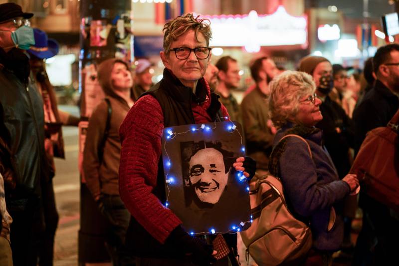A woman wearing glasses and a coat holds a picture of a man with lights on the border in a crowd of people.