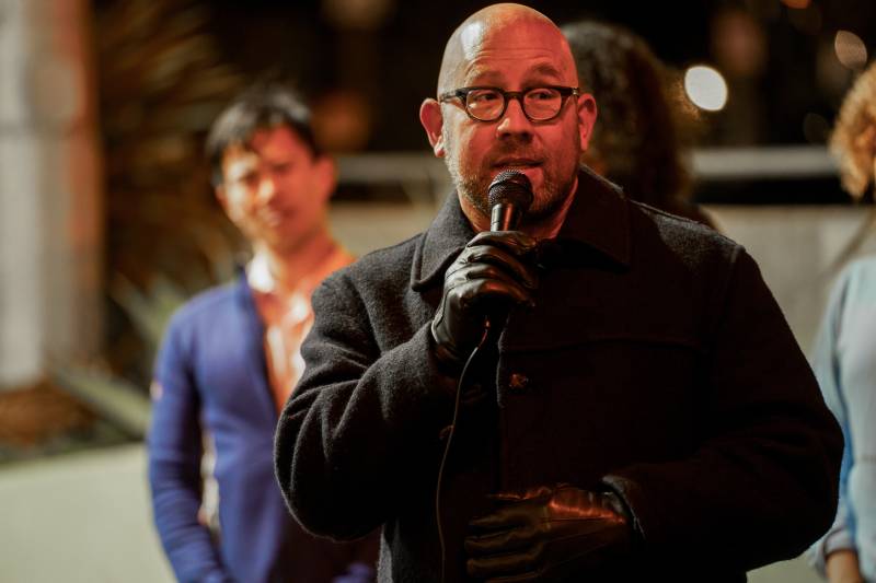A man wearing glasses and a coat hold a microphone outside around people.