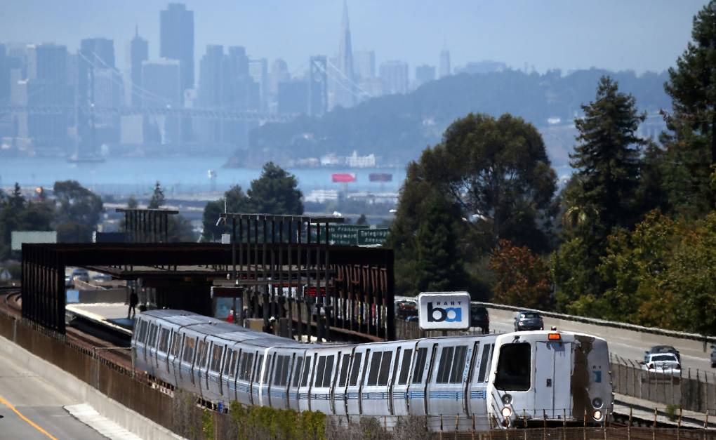 A BART train pulls out of an above ground station. Cars whiz by alongside and the city of San Francisco looms in the background.