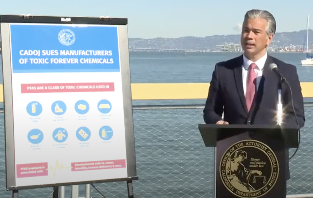 Rob Bonta speaking behind a dais with the Office of the Attorney General insignia, standing next to a board that shows the lawsuit his office filed, water and mountains in the background.