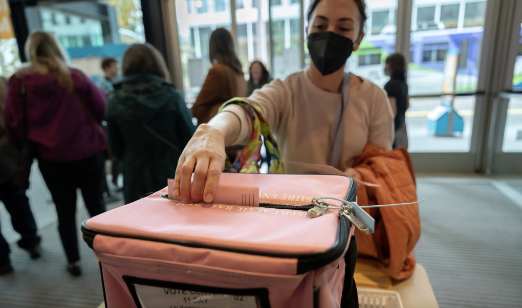 A woman wearing a black face mask places a piece of paper into a ballot box.