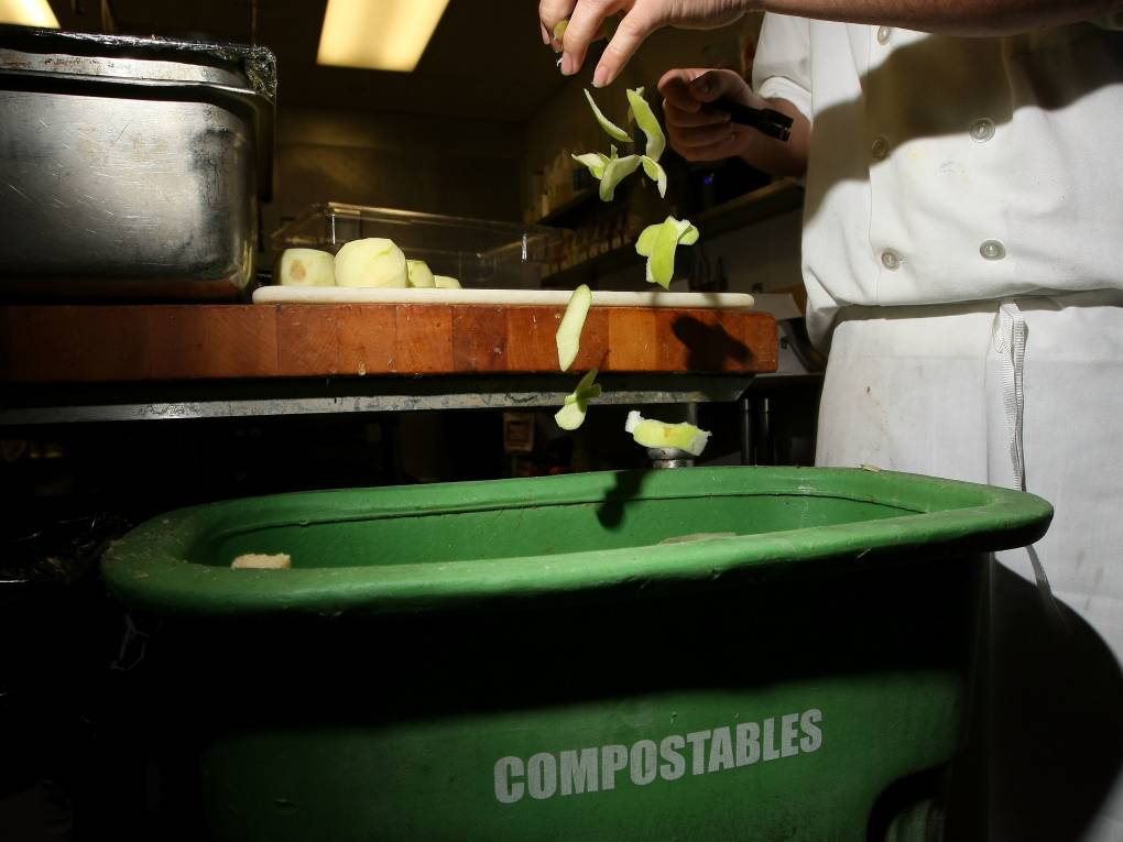 Food scraps from a chef chopping something drop into a green bin with the word 'compostable' on the side