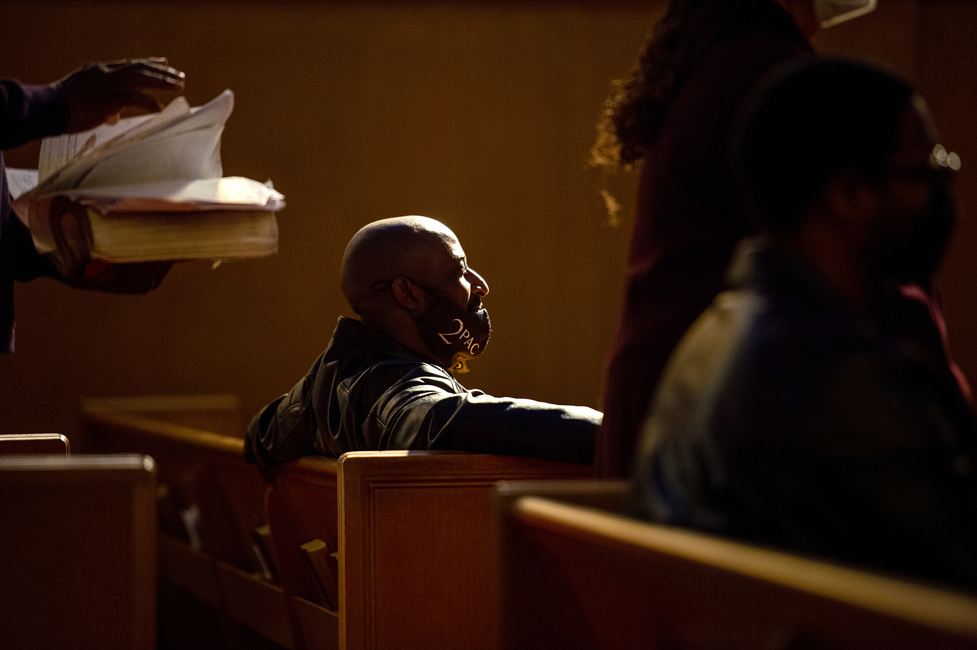 A Black man sits at a church pew, photographed in profile, looking like he is listening intently to something. He is wearing a cloth mask that is pulled down below his nose.
