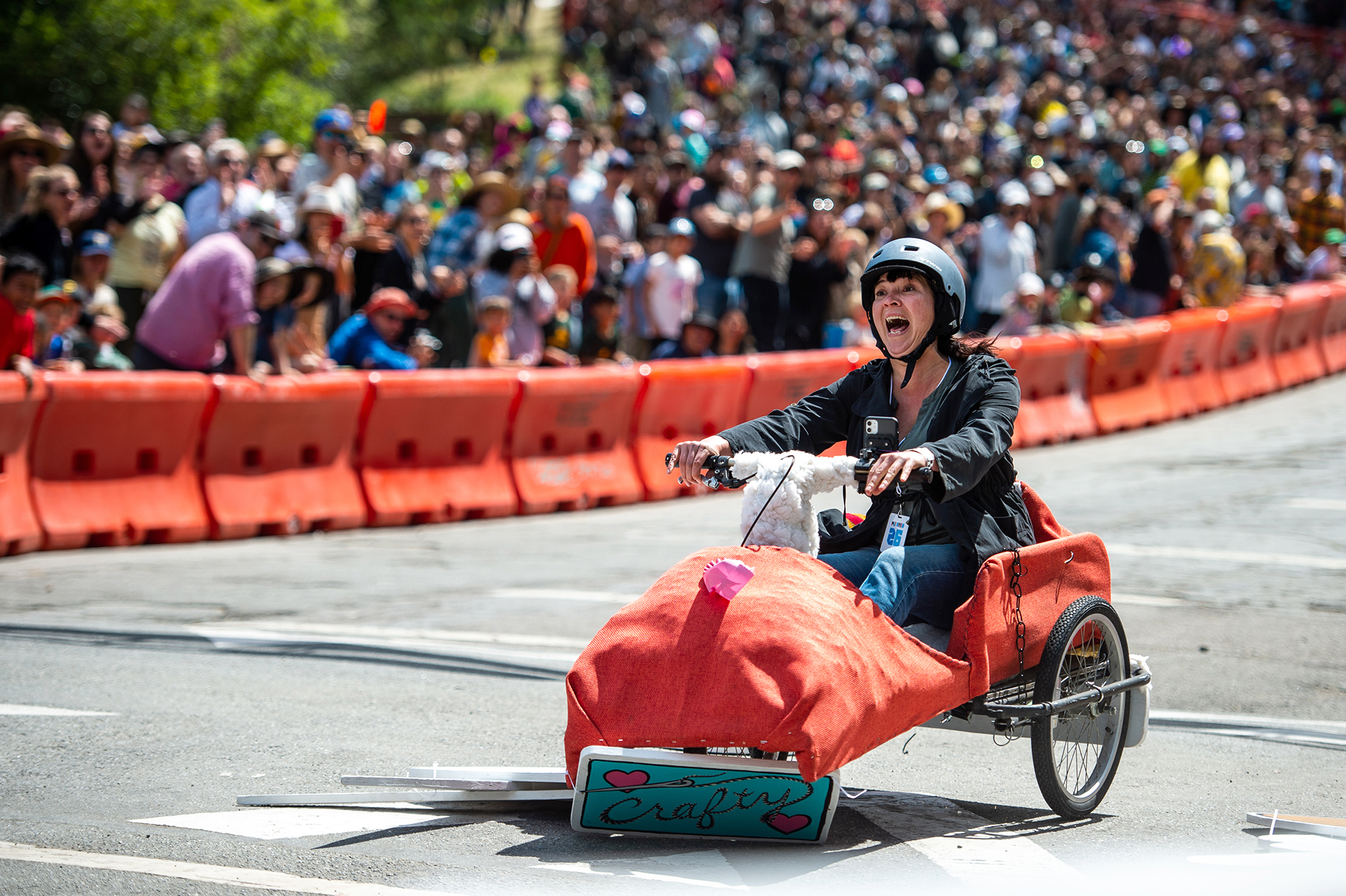 A person with light skin screams in what looks like a mix of excitement, delight and fear as they ride a red colored soapbox car down a hill. A crowd of people stands behind them out of focus behind a bright red barrier.