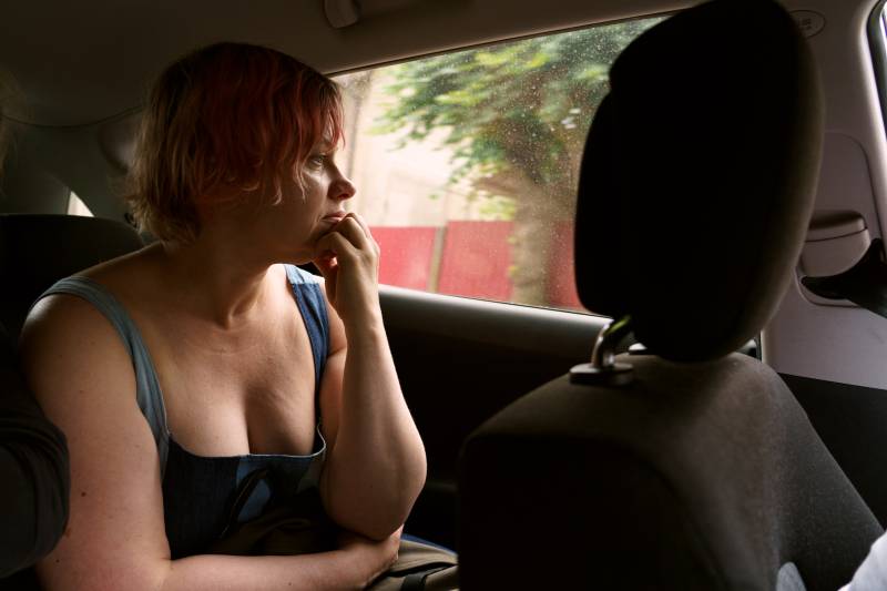 A youngish white woman with short, unkempt, light pink hair sits in the backseat of a car, the knuckles of her left hand holding her chin, looking out the window with a serious look on her face.